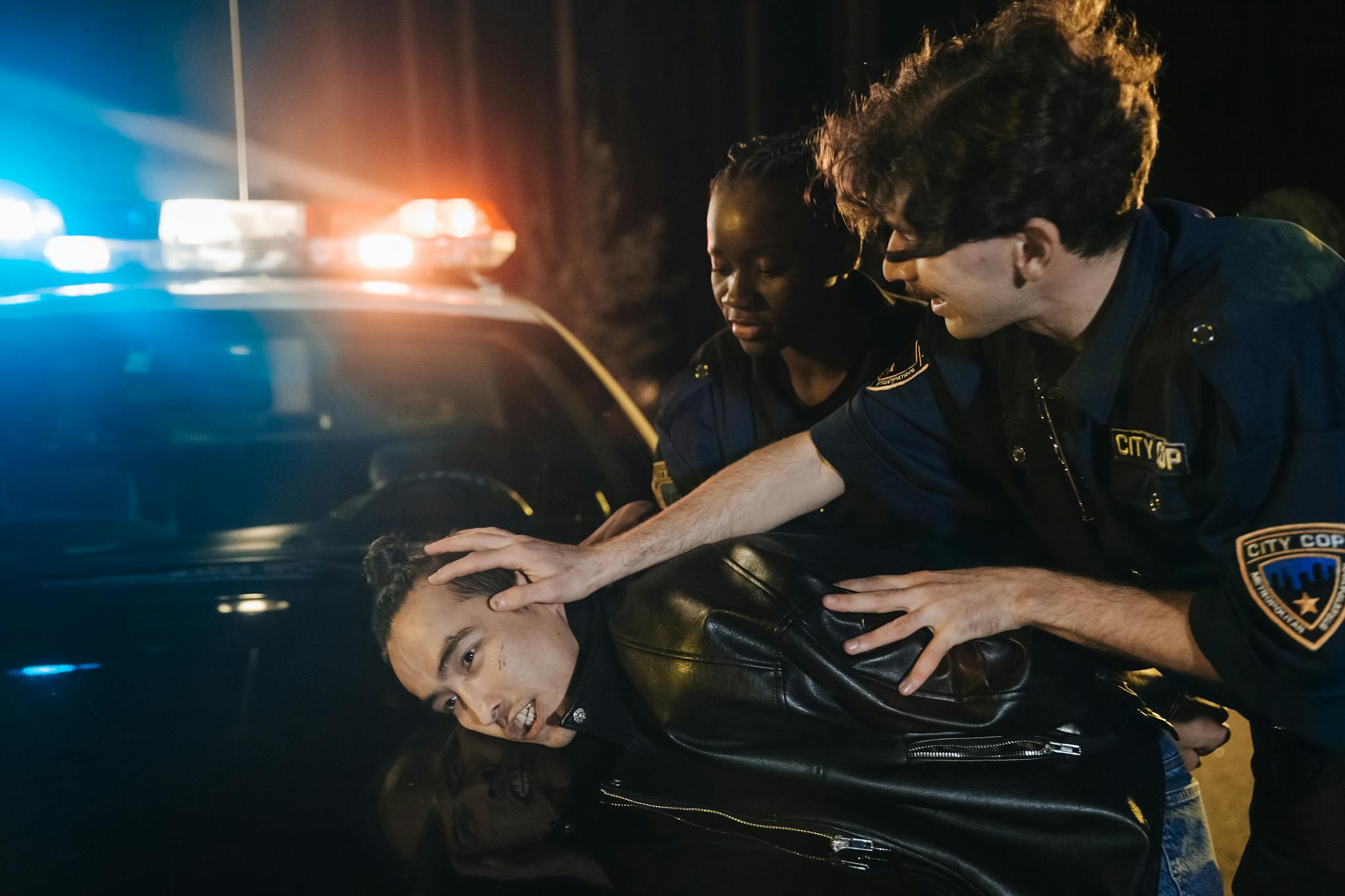 Two police officers arrest a suspect during a nighttime operation with flashing lights.